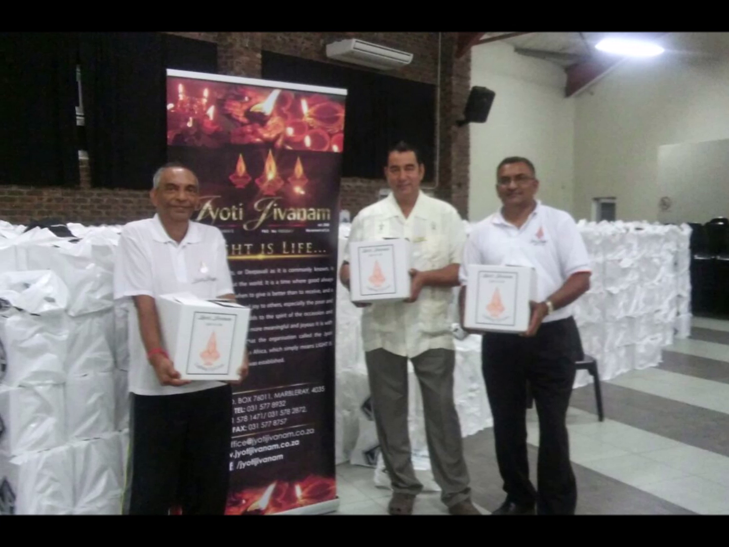 Three Jyoti Jivanam members presenting branded hamper boxes in front of distribution area