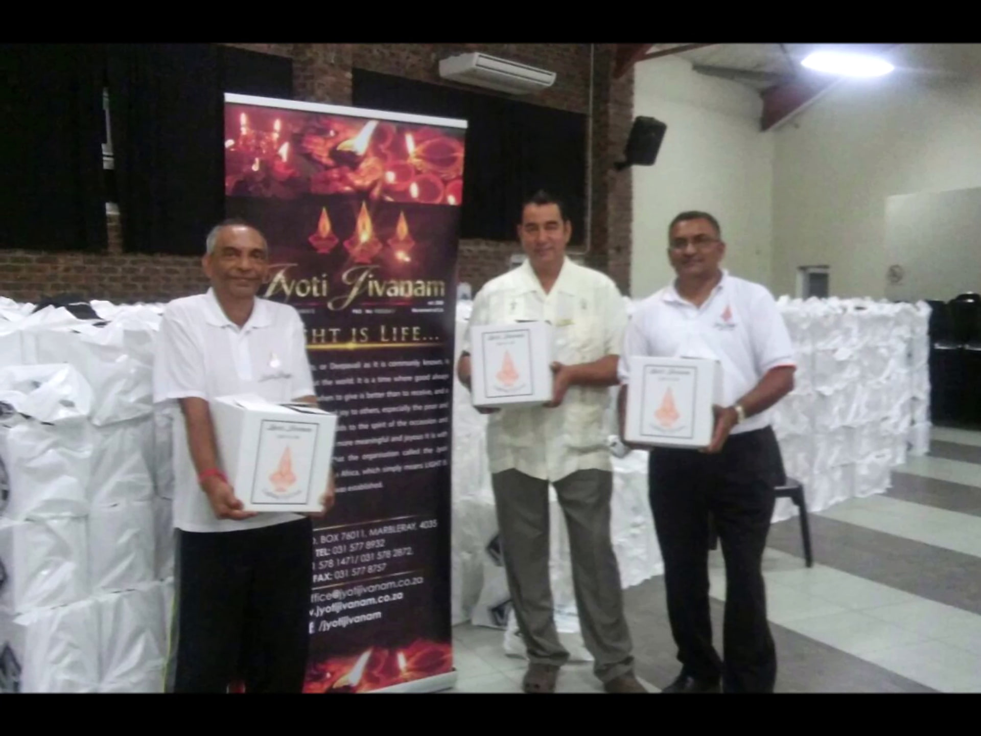 Three Jyoti Jivanam members presenting branded hamper boxes in front of distribution area